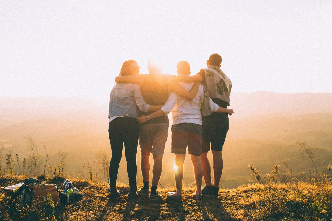 A group of friends standing together outdoors at sunset, embracing as they look out over the landscape.